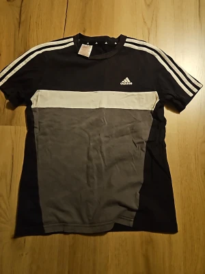 Svart och grå Adidas t-shirt - Snygg svart t-shirt från Adidas med grå panel framtill och vit rand över bröstet. Klassiska vita Adidas-ränder längs axlarna och logga på bröstet. Tillverkad i mjuk bomull och har korta ärmar. Perfekt för en sportig och avslappnad stil.