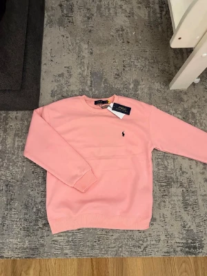 Rosa sweatshirt från Polo Ralph Lauren - Snygg rosa sweatshirt från Polo Ralph Lauren med klassisk rund halsringning och liten broderad logga på bröstet. Tröjan har ribbade muddar vid ärmslut och nederkant. Perfekt för en avslappnad och stilren look.