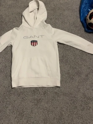 Vit hoodie från GANT med logga - Snygg vit hoodie från GANT med klassisk logga och sköld på bröstet. Hoodien har huva, magficka och ribbade muddar vid ärmslut och nederkant. Perfekt för en chill och stilren look. Materialet känns mjukt och bekvämt, troligen bomullsmix.