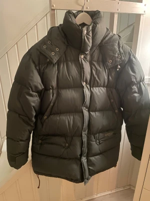 Mörkgrön pufferjacka från Everest - Säljer en mörkgrön pufferjacka från Everest med hög krage, avtagbar huva och dragkedja med tryckknappar. Jackan har två stora fickor framtill och quiltad design som ger en riktigt puffig look. Perfekt för kalla dagar och riktigt snygg till streetwear.