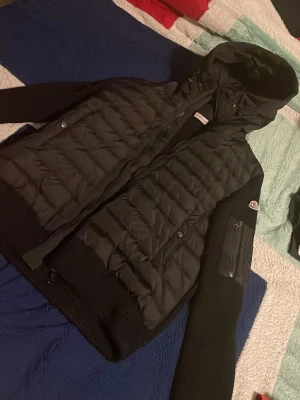 Svart dunjacka från Moncler med huva - Snygg svart dunjacka från Moncler med quiltad front och stickade ärmar. Jackan har huva, dragkedja framtill och en praktisk ficka med dragkedja på ärmen. Perfekt för kyliga dagar och riktigt stilren look.