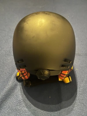 Svart hjälm från Anon - Säljer en svart hjälm från Anon med matt finish och ventilationshål. Hjälmen har öronlappar i röd och orange rutigt tyg samt gula spännen. Perfekt för vintersport och skydd på brädan eller i backen.