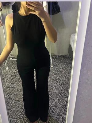 Svart jumpsuit med öppen rygg - Snygg svart jumpsuit med ärmlös design och öppen rygg. Vida ben och figurnära passform upptill. Perfekt för dig som vill ha en stilren och trendig look. Materialet känns mjukt och följsamt mot kroppen.