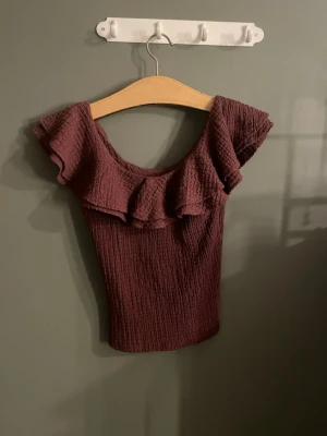 Vinröd offshoulder topp med volang - Trendig vinröd offshoulder topp med volangdetalj runt halsen. Tillverkad i ett strukturerat, stretchigt material som sitter snyggt och bekvämt. Perfekt för dig som vill ha en unik och stilren look.