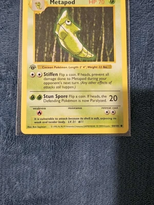 Metapod #54 1st Edition  - Säljer ett Metapod Pokémonkort från Base Set 1999. Kortet har gul ram, grön illustration av Metapod och detaljer om attacker och stats. Baksidan har klassisk blå design med Pokéball. Perfekt för samlare eller Pokémonfans!