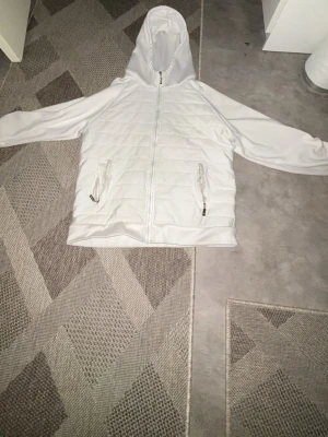 Vit vindjacka med huva och dragkedja - Snygg vit vindjacka med huva och hel dragkedja framtill. Jackan har två fickor med dragkedja och quiltade detaljer på framsidan. Insidan är fodrad med blankt tyg för extra komfort. Perfekt för blåsiga dagar och den är storlek M.