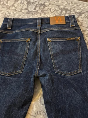 Mörkblå selvedge jeans från Nudie Jeans 29/32 - Snygga mörkblå jeans från Nudie Jeans med klassisk femficksdesign och kontrastsömmar. Jeansen har en rak passform och är tillverkade i slitstarkt denimtyg av bomull. Perfekta för dig som gillar stilrena och tidlösa jeans.
