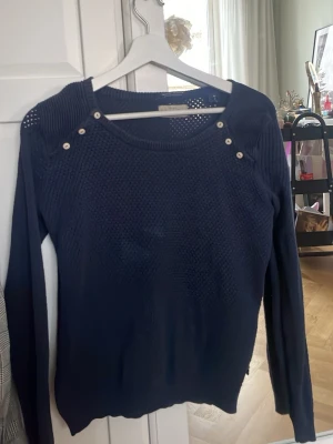 Mörkblå stickad tröja med knappar - Mörkblå stickad tröja med rund halsringning och dekorativa vita knappar på axlarna. Tröjan har lång ärm och ett diskret strukturmönster framtill. Perfekt att matcha med jeans eller kjol för en snygg vardagslook.