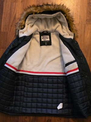 Svart parkas från Stone Goose - Snygg svart parkas från Stone Goose med fuskpäls på huvan och quiltade detaljer på axlar och insida. Jackan har dragkedja och knappar framtill, flera fickor och ett varmt, mjukt innerfoder i vitt med röda och svarta ränder. Perfekt för kalla dagar.