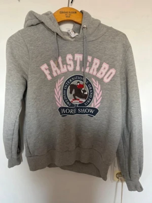 Grå Falsterbo hoodie med tryck - Snygg grå hoodie från Falsterbo med huva och dragsko. Stor tryckt logga på bröstet med texten 'Falsterbo Horse Show' i rosa och blått samt motiv av ryttare. Mjuk bomullsblandning och ribbade muddar vid ärmar och midja. Perfekt för dig som gillar hästsport och vill ha en sportig look. Obs! En del använd och nopprig på insidan och defekt vid kragen
