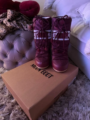 Moonboots - Vinröda Moon Boot med vit logga och snörning framtill. Klassisk rund form och platt sula, tillverkade i syntetmaterial med vadderad känsla. Perfekta för vintern. Hej! Säljer mina moonboots dom har använts max 4 gånger och är i toppen skick