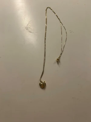 Guldigt hjärthalsband från Pilgrim - Snyggt halsband från Pilgrim med ett hjärtformat hänge i guldton. Kedjan är tunn och elegant, perfekt för att ge en söt touch till din outfit. Hjärtat har en blank finish och Pilgrim-loggan ingraverad på framsidan. Kedjan är även justerbar.
