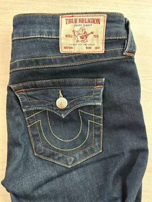 True Religion mörkblå jeans - Mörkblå jeans från True Religion med ikonisk bakficka och kontrastsömmar i gult och orange. Klassisk femficksmodell med tydlig logga på bakfickan och patch i läderimitation vid linningen. Perfekta för dig som gillar streetwear och snygga detaljer. Säljer för att de är lite för långa och har inte kommit till användning.