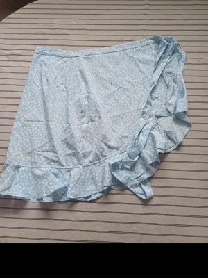 Blommig blå omlottkjol Vero Moda - Söt ljusblå omlottkjol från Vero Moda med vitt blommönster och volangkant. Kjolen är kort och har en luftig känsla, perfekt för varma dagar. Tillverkad i mjuk polyester som känns skön mot huden.