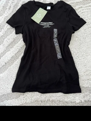 Svart ribbad t-shirt från H&M - Snygg svart ribbad t-shirt från H&M med trycket 'FUTURISTIC FEELS' och text framtill. Tillverkad av 25% återvunnen bomull. Klassisk rund hals och korta ärmar, perfekt för en trendig och avslappnad stil.