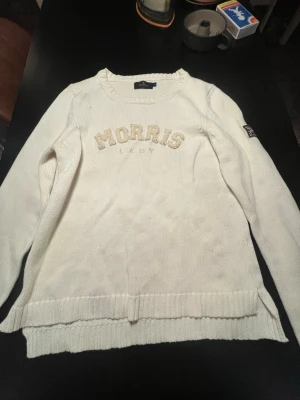 Vit stickad tröja från Morris Lady - Snygg vit stickad tröja från Morris Lady med broderad logga framtill och Union Jack-detalj på ärmen. Tröjan har rund halsringning, långa ärmar och ribbade muddar. Perfekt för dig som gillar klassisk och stilren design.