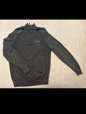 Grå halvzip-tröja från Hugo Boss - Snygg grå tröja från Hugo Boss med halv dragkedja och blå detaljer på axlarna. Broderad logga i blått på bröstet. Långärmad modell med ribbade muddar och stilren passform. Perfekt för en trendig och avslappnad look.