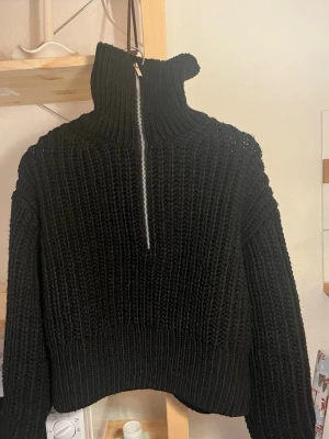 Svart stickad tröja med halvzip - Säljer en svart stickad tröja med hög krage och silverfärgad dragkedja framtill. Tröjan har en chunky ribbstickad struktur, långa ärmar och avslappnad passform. Perfekt för kalla dagar och enkel att styla till olika outfits.