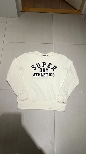 Vit sweatshirt från Superdry - Vit sweatshirt från Superdry med svart broderad text 'SUPERDRY ATHLETICS' på bröstet. Klassisk rund halsringning och ribbade muddar vid ärmslut och nederkant. Perfekt för en sportig och avslappnad stil.