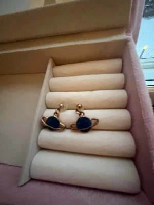 Guldiga örhängen med planetmotiv - Säljer ett par coola örhängen i guldmetall med blå planetformad detalj. De har en unik design som liknar Saturnus med ringar runt planeten. Perfekta för dig som gillar rymdinspirerade accessoarer.🚀💙