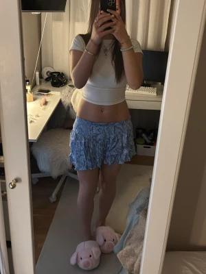 Blå volangkjol med blommigt mönster - Säljer en söt blå kortkjol med volanger och blommigt mönster. Kjolen har en luftig och flowy känsla, perfekt för varma dagar. Den har resår i midjan för en bekväm passform och är lätt att matcha med olika toppar. Helt oanvänd