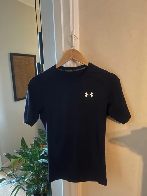 Svart träningströja från Under Armour - Snygg svart träningströja från Under Armour med korta ärmar och rund hals. Tröjan har en diskret logga på bröstet och är tillverkad i ett lätt och snabbtorkande material som passar perfekt till gymmet eller löprundan.