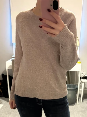 Beige stickad tröja i enkel design - Mjuk och stilren beige stickad tröja med rund halsringning och långa ärmar. Tröjan har ribbade muddar vid ärmslut och nederkant, perfekt för lager på lager. Passar till både jeans och kjol för en avslappnad look.