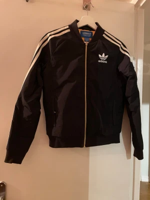 Svart Adidas bomberjacka med vita ränder - Snygg svart bomberjacka från Adidas med klassiska vita ränder längs axlarna och ärmarna. Jackan har dragkedja framtill, ribbade muddar och Adidas logga på bröstet. Perfekt för en sportig och trendig look.