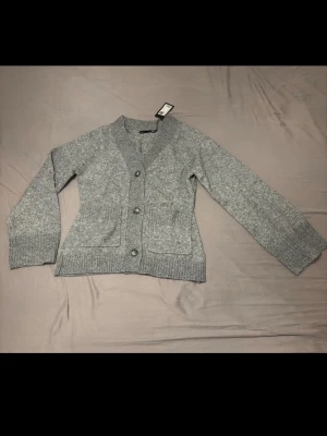 Grå stickad kofta med fickor - Mysig grå stickad kofta med v-ringning och tre knappar framtill. Koftan har två stora fickor och ribbstickade muddar vid ärmslut och nederkant. Storlek XS. Helt ny från lager 157, aldrig använd och med prislapp kvar!!!🌸