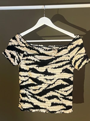 Zebramönstrad smockad topp - Trendig zebramönstrad topp med smockad struktur och korta ärmar. Toppen har en off shoulder-modell och vågig kant nertill och vid ärmslut. Färgerna är svart och beige, vilket ger en cool kontrast. Perfekt för dig som gillar djurmönster och vill sticka ut. Köpt från JDY / Nelly, aldrig använt lappen sitter kvar 🦓🤍