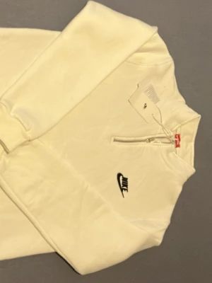 Vit Nike hoodie med halv dragkedja - Snygg vit hoodie från Nike med halv dragkedja framtill och svart Nike-logga broderad på bröstet. Tröjan har ribbade muddar vid ärmslut och nederkant samt en klassisk, stilren design. Perfekt för dig som gillar sportig och clean streetwear.