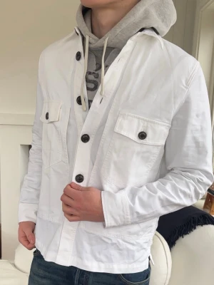 Vit overshirt med knappar Jacob Cohën  - Stilren vit overshirt med stora svarta knappar och två bröstfickor med lock. Långärmad modell i bomull som passar perfekt över en hoodie för en avslappnad look. Snygg detalj i nacken och klassisk passform.