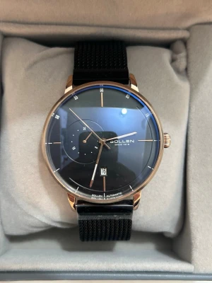 Svart och guld klocka från Sollen - Stilren klocka från Sollen med svart urtavla, guldfärgade detaljer och svart mesh-armband. Klockan har datumvisning och en extra liten 24-timmarsvisare. Boetten är rund och elegant, perfekt för dig som gillar minimalistisk design.