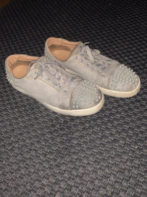 Grå Christian Louboutin skor  - Säljer ett par grå Christian Louboutins  i mocka med coola spikar på tån och hälen. Skorna har snörning och en klassisk platt sula. Insidan är beige och designen är riktigt edgy och unik. Perfekta för dig som vill sticka ut med din stil. Jätte feta men tyvärr saknar ett par spikar därav priset! 