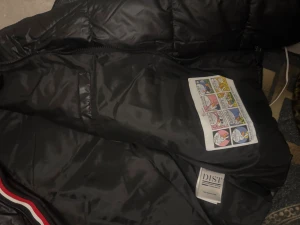 Svart Moncler pufferjacka med huva - Svart pufferjacka från Moncler med huva och coolt Moncler-märke på bröstet. Jackan har en röd, vit och blå rand längs dragkedjan och quiltad design. Insidan har en unik serietidningsetikett. Perfekt för dig som vill ha en snygg och varm jacka med street vibe.