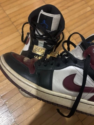 Nike Air Jordan 1 High svart/vinröd - Nike Air Jordan 1 High sneakers i svart, vinröd och vit med klassisk rund tå och svarta snören. Skorna har guldfärgade detaljer på snörningen med text som '1985', 'JORDAN' och 'WOMEN'. Tillverkade i skinn och syntetmaterial.