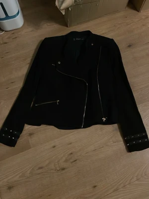 Svart skinnjacka från Zara Trafaluc - Snygg svart skinnjacka från Zara Trafaluc med asymmetrisk dragkedja och detaljerade ärmslut med spännen. Jackan har en modern, kortare passform och flera dragkedjor framtill som ger en cool look.