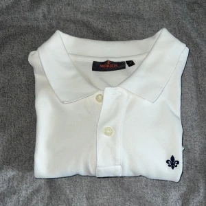 Perfekt Vit pikétröja från Morris - Vit pikétröja från Morris med klassisk krage och två knappar framtill. Liten broderad logga i form av en mörkblå lilja på bröstet. Perfekt basic-plagg med stilren design. Fungerar perfekt nu till sommaren och är praktiskt taget oanvänd typ. Storlek M och true to size. 