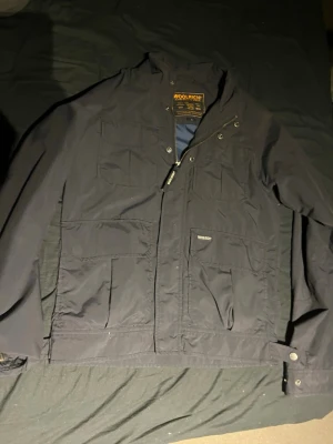 Svart vindjacka från Woolrich - Svart vindjacka från Woolrich med flera praktiska fickor framtill och dragkedja. Jackan har en klassisk passform och är tillverkad i slitstarkt syntetmaterial. Perfekt för blåsiga dagar och enkel att matcha med olika outfits.