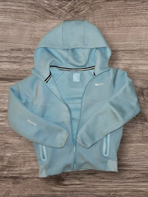 Ljusblå Nike NOCTA hoodie - Snygg ljusblå hoodie från Nike och NOCTA-kollektionen med dragkedja framtill, huva och vita detaljer. Broderad Nike-logga på bröstet och NOCTA-tryck på ärmen. Mjukt material och två fickor med dragkedja. Perfekt för en avslappnad streetwear-look.
