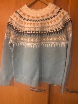Ljusblå stickad tröja med mönster - Mysig ljusblå stickad tröja med rund hals och klassiskt nordiskt mönster i vitt, beige och brunt upptill. Tröjan har långa ärmar och ribbade muddar vid ärmslut och nederkant. Perfekt för kalla dagar och ger en avslappnad men snygg look.