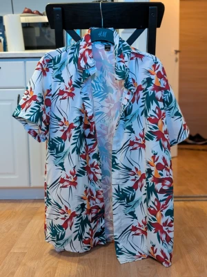 Kortärmad skjorta – tropiskt mönster – SHEIN – stl L – NY - Säljer en kortärmad skjorta med tropiskt/blommigt mönster från SHEIN. Helt oanvänd, köpt men blev för liten. 🔸 Storlek: En mindre storlek av L 🔸 Lätt och luftig – perfekt till sommaren 🔸 Kort ärm, knäppning framtill 🔸 Inga skador eller slitage Pris: 100 kr Kan skickas mot frakt eller hämtas upp. Hör av dig vid intresse!