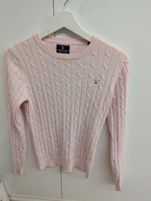 Ljusrosa kabelstickad tröja Gant - Supersöt ljusrosa kabelstickad tröja från Gant med rund halsringning och diskret logga broderad på bröstet. Tröjan har långa ärmar och ribbade muddar vid ärmslut och nederkant. Perfekt för dig som gillar klassisk och stilren look.                                                Pris kan diskuteras vid smidigt köp 😊.                        Köparen står för frakt!