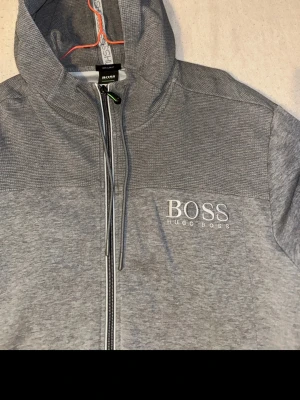 Grå hoodie från Hugo Boss - Snygg grå hoodie från Hugo Boss med dragkedja framtill och broderad logga på bröstet. Hoodien har justerbar huva med snören och två fickor på sidorna. Tillverkad i mjukt och bekvämt material, perfekt för en avslappnad stil.
