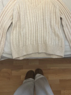 Beige ribbstickad tröja V-ringad - Säljer en beige ribbstickad tröja med V-ringning och långa ärmar. Tröjan har breda ribbor och en avslappnad passform, perfekt för lager på lager. Materialet känns mjukt och mysigt, och färgen är lätt att matcha med det mesta.