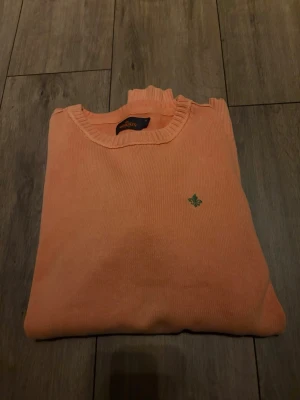 Persikofärgad stickad tröja Morris - Persikofärgad stickad tröja från Morris med rund halsringning och ribbade muddar. Tröjan har en liten grön broderad logga på bröstet och är tillverkad i ett mjukt material. Perfekt för dig som vill ha en stilren och enkel look.