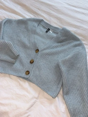 Ljusblå cropped kofta med knappar - Superfin ljusblå cropped kofta med tre stora bruna knappar framtill. Koftan har v-ringning, lång ärm och är stickad i ett mjukt material.