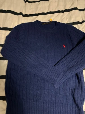 Mörkblå kabelstickad tröja Polo Ralph Lauren - Kabelstickad mörkblå tröja från Polo Ralph Lauren med rund halsringning och lång ärm. Tröjan har det klassiska röda Polo-logobroderiet på bröstet och ribbade muddar vid ärmslut och nederkant. Perfekt för dig som gillar stilren och tidlös design.(den har krympt lite så hade sagt att den passar en S person)