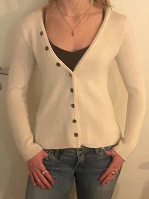 Beige kofta med sned knäppning - Stilren beige kofta med lång ärm och unik sned knäppning med svarta knappar. Tunnstickad modell med v-ringning, perfekt att bära över ett linne eller t-shirt. Enkel och trendig design som passar till jeans eller kjol. Pris kan diskuteras!🫰🏻