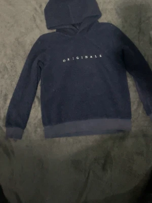 Mörkblå hoodie Originals - Mysig mörkblå hoodie från Originals med tryckt text på bröstet. Tröjan har huva, långa ärmar och ribbade muddar vid ärmslut och nederkant. Materialet ser ut att vara fleece eller teddy, perfekt för en avslappnad stil.